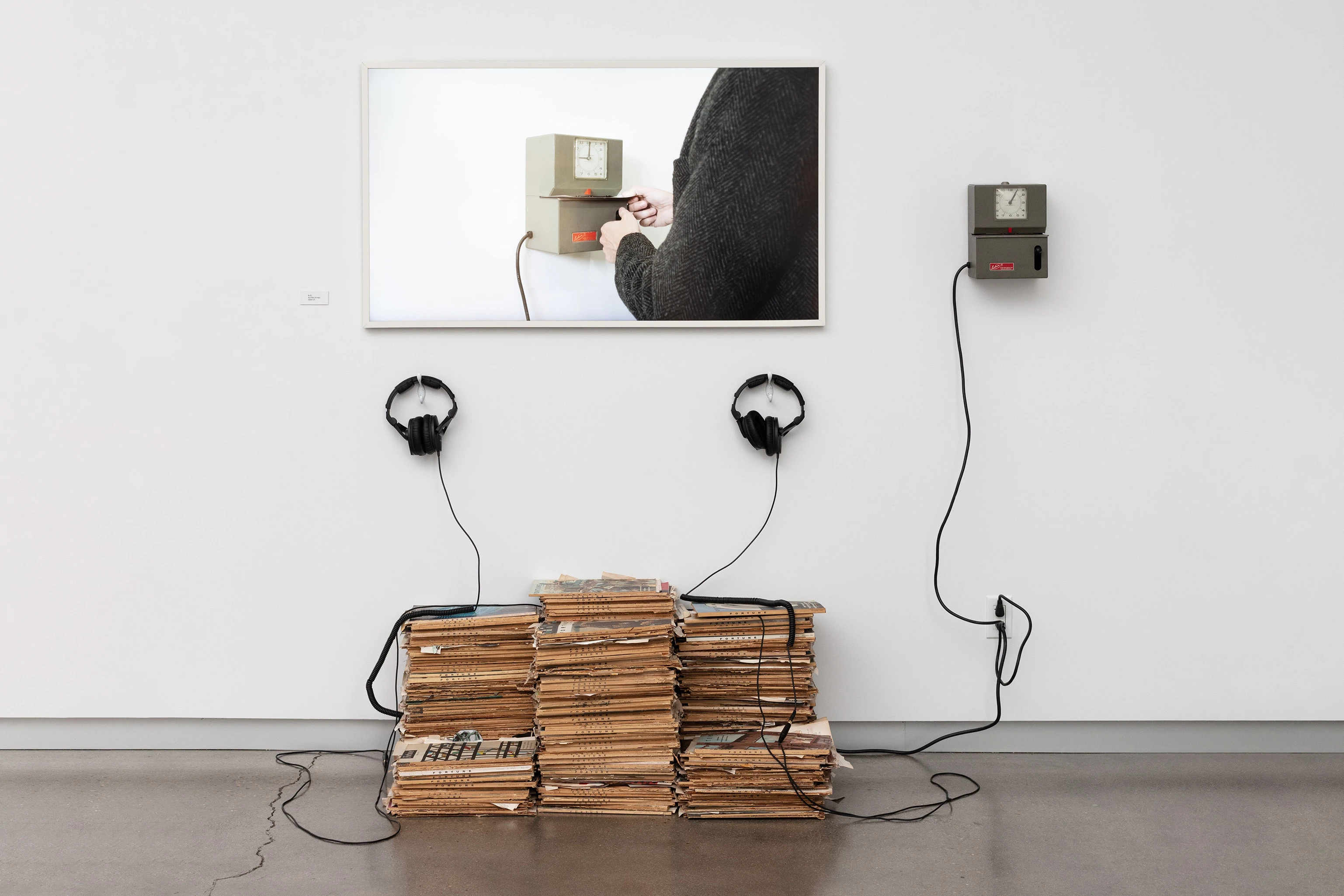 Installation view of screen, stacks of magazine, headphones, and punch-in clock against a gallery wall